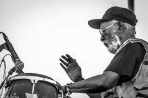 Black-and-white photo of a man in a cap playing a drum, with a microphone visible on the left.