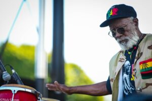 Older man with gray beard wearing a colorful shirt and cap, playing a red conga drum on an outdoor stage beside a microphone.