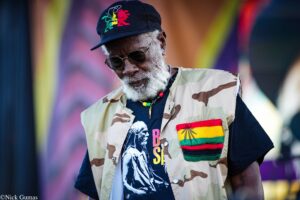 Older man with a white beard wearing a camouflage vest, black shirt, round sunglasses, and a cap with a rainbow lion motif against a colorful blurred background.