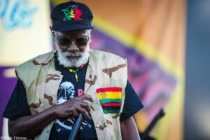 Older man with a white beard wearing a camouflage vest, a black cap with a colorful lion design, sunglasses, and beaded necklace, holding a microphone at a performance.