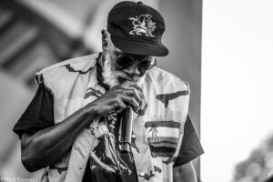 Black-and-white photo of an older man singing into a microphone, wearing a camouflage vest and cap with an emblem, plus sunglasses.