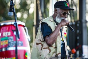 Older man with gray beard wearing a camo vest and dark sunglasses sings into a handheld microphone at an outdoor performance.