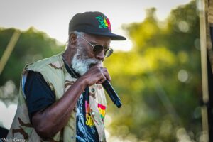 Older man with a gray beard singing into a microphone outdoors, wearing a camouflage vest and black cap with colorful embroidery, sunglasses, and a crowd- or park-like background.