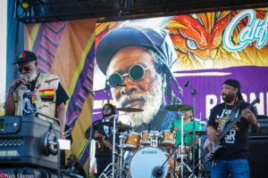 Musicians perform on a colorful stage, with a drummer center, guitarist right, and a vocalist in a camo vest and cap in the foreground.
