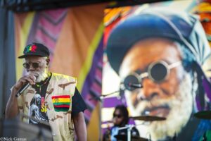 Male singer wearing a camo vest and black cap, gripping a microphone on a colorful mural-backed stage.