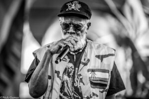 Black-and-white photo of an elderly man wearing a cap and round glasses, singing into a microphone while performing.