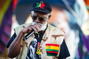 Older man with a white beard wearing sunglasses and a camouflage vest, singing into a handheld microphone on stage.