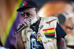Older man with a gray beard singing into a microphone, wearing a black cap and round sunglasses with a camouflage vest.