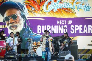 Live band on a colorful festival stage; lead singer in camo with microphone, guitarists and drummer in the background, bold Backdrop reads 'Next Up Burning Spear' at California Roots 2026.