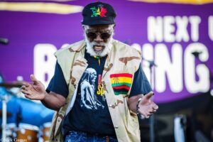 Older man with a gray beard wearing a camouflage vest, black cap with a colorful emblem, and sunglasses performing on a purple-stage backdrop.