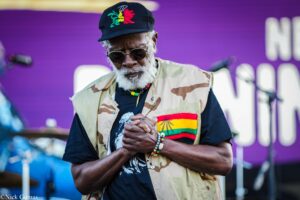 Older man with gray beard and sunglasses, wearing a camouflage vest and colorful beaded bracelets, hands clasped on stage in front of a purple backdrop.