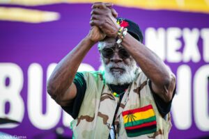 Older man with gray beard and sunglasses, in camouflage clothing and Rastafari-colored sash, raises clasped hands in front of a purple banner.