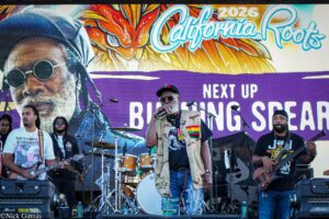 Live band performs on a sunny stage at California Roots 2026, with a lead singer in a camouflage shirt and cap, guitarist on the left, drummer behind, and a colorful backdrop behind them reading 'California Roots 2026' and 'Next Up'.
