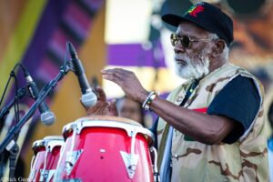 Older man with a gray beard wearing a camo vest and black cap plays a red conga drum on stage, microphone in front of him.