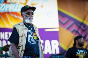 Older man with a gray beard and round sunglasses, wearing a black cap and camouflage vest at a colorful outdoor festival backdrop.