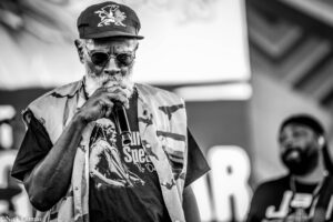 Older man with a beard wearing sunglasses, a camo jacket and a cap, singing into a microphone at a performance (black and white).