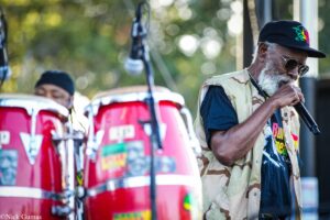 Older man with a gray beard wearing sunglasses and a camouflage vest, singing into a microphone on stage beside red drums outdoors.