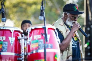 Older man with gray beard wearing a navy cap and sunglasses sings into a microphone at an outdoor performance, red drums in foreground.
