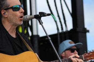 Male guitarist sings into a microphone on stage, wearing sunglasses and an in-ear monitor with an acoustic guitar close in view