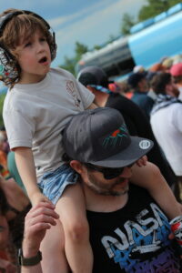 Child on a man's shoulders wearing large headphones at an outdoor event, with a blue bus and crowd in the background