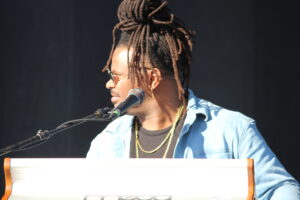 Musician with brown dreadlocks singing into a microphone while playing a white keyboard on stage. (side profile)