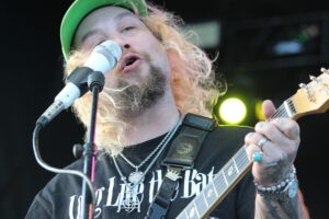 Guitarist with long blond hair and beard singing into a microphone on stage, wearing a green cap and black T-shirt.