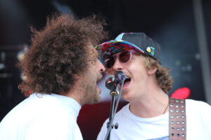 Two male singers share a microphone on stage during a live performance; one with curly hair in a white shirt and the other wearing a colorful cap and pink sunglasses.