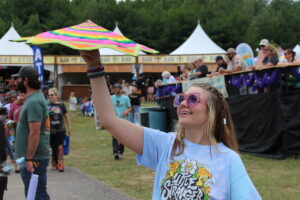 Girl in a light blue shirt raises a colorful rainbow kite at a crowded festival with white tents in the background.