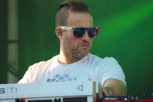 Male musician wearing clear-framed sunglasses and a white t-shirt, playing a keyboard on a green-lit stage, in-ear monitors visible on ear.