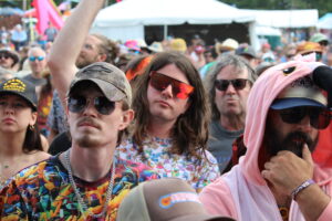 Crowd at an outdoor festival; people wearing bright clothing and sunglasses, packed together near tents and booths.