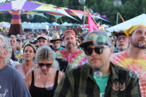 Crowd of festival-goers in tie-dye shirts at an outdoor event with colorful tents in the background.