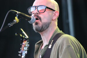 Bald male singer with a beard and sunglasses singing into a microphone on stage, guitar strap across his chest.