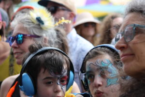 Close-up of a child with blue headphones and face paint among a colorful crowd at an outdoor event.