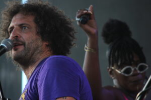 Male singer with curly hair wearing a purple shirt, singing into a microphone on stage, raised hand in background.