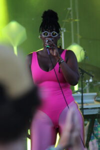 Female singer in a bright pink sleeveless outfit performs on stage, wearing jeweled round sunglasses and holding a microphone.