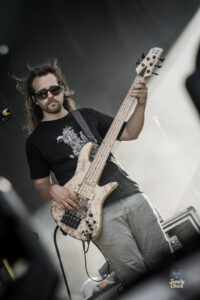 Male bass guitarist with long hair and sunglasses playing a cream-colored bass on stage during a live performance.