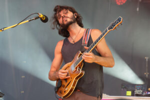 Male guitarist performing on stage with a striped strap, wearing a black tank top and playing an orange-brown electric guitar near a microphone.