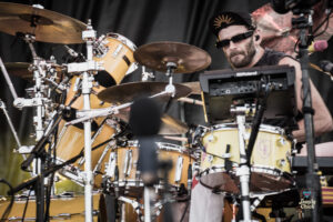 Drummer with sunglasses and a black cap performing on a yellow drum kit with multiple cymbals on stage.