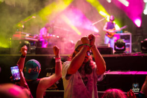 Front-row concert scene with hands raised in the air, colorful stage lights, and a guitarist performing on a smoky stage.