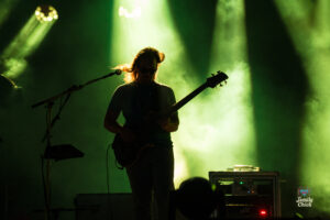 Silhouetted guitarist on stage under green spotlight with fog and audio gear visible, performing live.