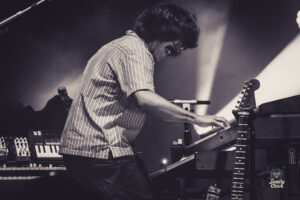 Musician in a striped shirt playing keyboards while bending toward a guitar on a dimly lit stage with electronic gear nearby.