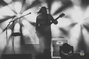 Bassist playing a guitar on stage with bright spotlights and smoky atmosphere in a black-and-white concert photo.