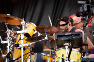 Drummer in sunglasses and a black cap playing a yellow drum kit on stage, with cymbals in the foreground.
