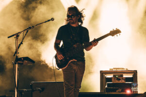 Bass guitarist performing on a smoky stage with warm yellow light, standing beside amplifiers and mic stand.