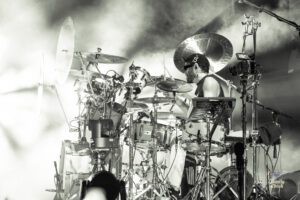 Drummer at a large drum kit on a smoky stage, surrounded by cymbals and microphones.