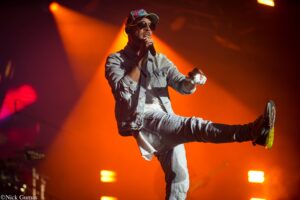 Male performer wearing a denim jacket and cap sings into a mic while mid‑jump on a brightly lit concert stage with orange lights.