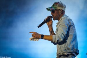 Male singer wearing a colorful floral cap and sunglasses, singing into a handheld microphone on a blue-lit stage background.