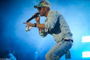 Male singer performing on stage, wearing a denim jacket and jeans, floral cap, sunglasses, and wireless earpiece while holding a microphone with a towel in hand against a blue background