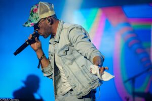 Male singer wearing a floral cap and denim jacket, singing into a microphone on a brightly lit blue stage with colorful lights.