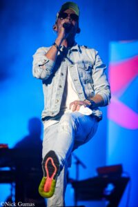 Male singer on stage mid-jump, wearing denim jacket, cap, sunglasses, and neon sneakers under blue stage lights, mic in hand.